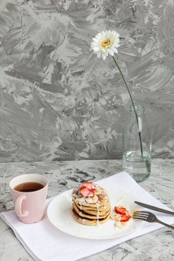 Arka planda beyaz bir tabakta taze çilekli krep yığını. Cam vazoda beyaz bir çiçek kahvaltıda yanında duruyor.. 