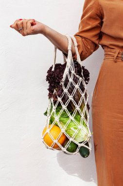 Kız elinde sebzeli örgü alışveriş poşeti, poşetsiz yeşillikler tutuyor. Sıfır atık, plastiksiz çevre dostu kavram. El yapımı makrome çanta. Sürdürülebilir yaşam tarzı.  