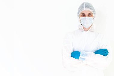 Koruyucu takım elbiseli ve yüz maskeli bir adam kollarını kavuşturmuş kameraya bakıyor. Beyaz bir arkaplanda izole edilmiş.