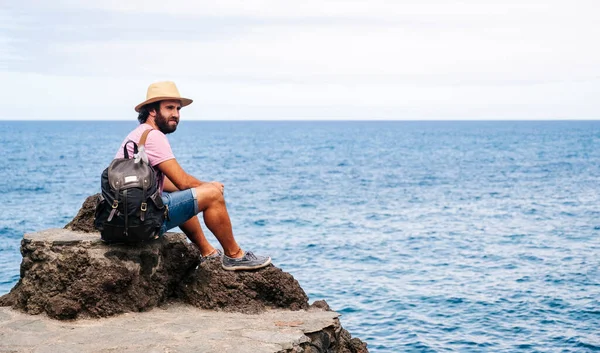 Arka planda deniz olan, sırt çantalı ve şapkalı bir turist..