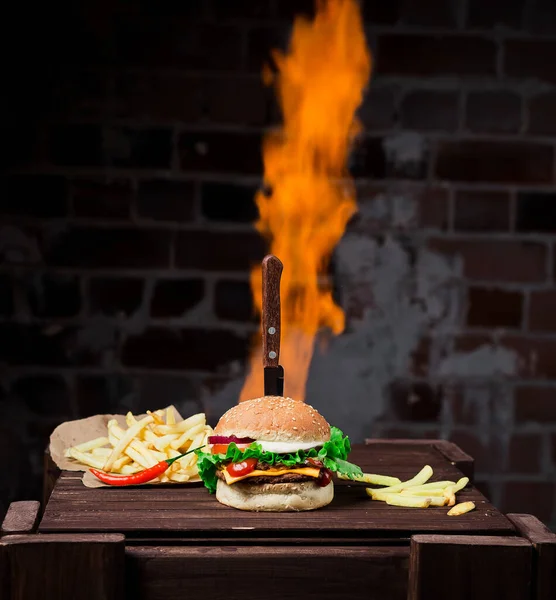 Stuck knife in a hamburger with French fries, a brown wooden table and a flame in the background, shot in studio.brick wall