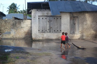 İki çocuk bir köy caddesinde yalınayak geziyor. Yağmur sonrası su birikintilerinde. Çocuklar basit oyuncaklarını yanlarında sürüklerler.