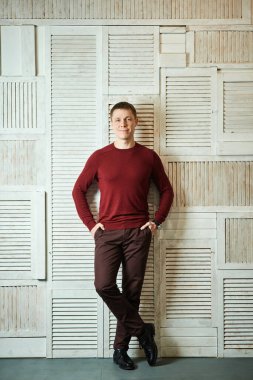 Yakışıklı genç adam, ayakları çaprazlanmış, sırtı duvara yaslanmış ellerini cebine sokmuş, gülümsüyor, kameraya bakıyor..