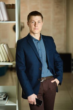 Şık genç adam, omzunu rafa dayayıp telefonu elinde tutuyor ve kameraya bakıyor..