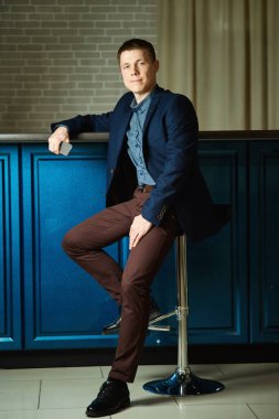 Genç bir adam bir bar taburesinde oturuyor, büyürken, tezgaha yaslanıyor, cep telefonu elinde..