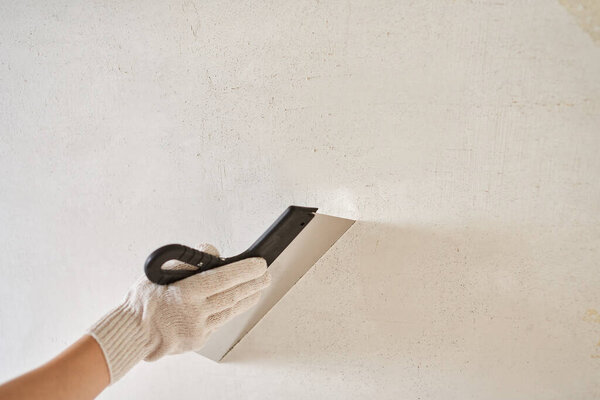 Spatula with putty in hand with a glove against a light wall. Worker puts of plaster on wall. Repair work. Plastering wall. Close-up.