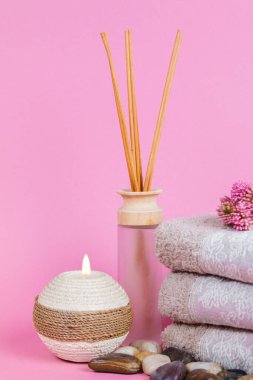 Spa malzemeleri, aroma çubukları taşlar, havlular ve pembe arka planda bir bitki.