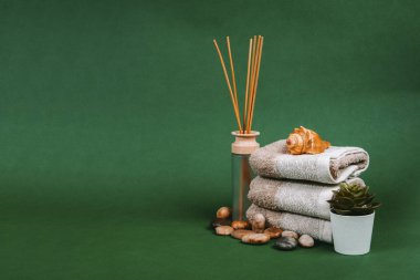 Kaplıca malzemeleri, mum, taşlar, havlu ve çiçekler koyu yeşil arka planda.