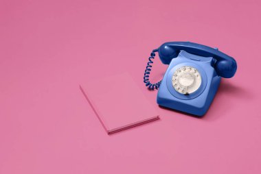 Pembe arka planda, fotokopi alanı ve metin için odası olan pembe bir defterin yanındaki antika mavi döner telefon.