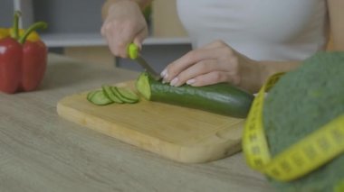 Beyaz bir kadın masada oturur ve bir salatalığı doğrar. Çerçeveyi kapat