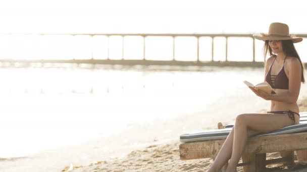 A menina senta-se em uma cadeira de praia junto ao mar e lê um livro — Vídeo de Stock