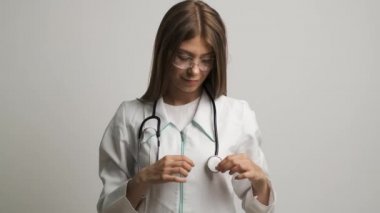 Beyaz önlüklü ve steteskoplu gülümseyen genç bayan doktor beyaz kardiyolog kameraya bakarak kalp şeklini gösteriyor.