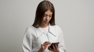 Beyaz önlüklü, beyaz önlüklü bir kız doktor telefona bakar ve kameraya gülümser.