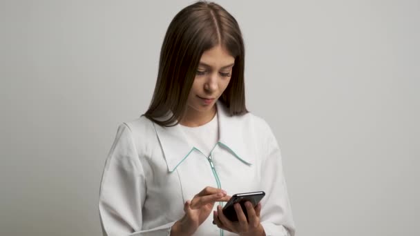 Menina médica em um casaco branco de aparência caucasiana olha para o telefone e sorri para a câmera — Vídeo de Stock