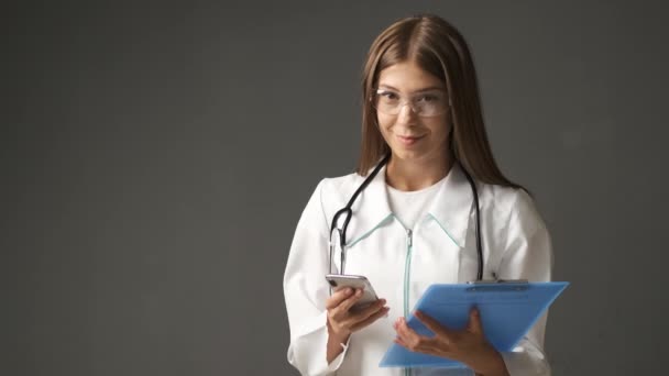 Ragazza medico in un cappotto bianco di aspetto caucasico tiene un telefono e una cartella con documenti sorridenti alla fotocamera — Video Stock