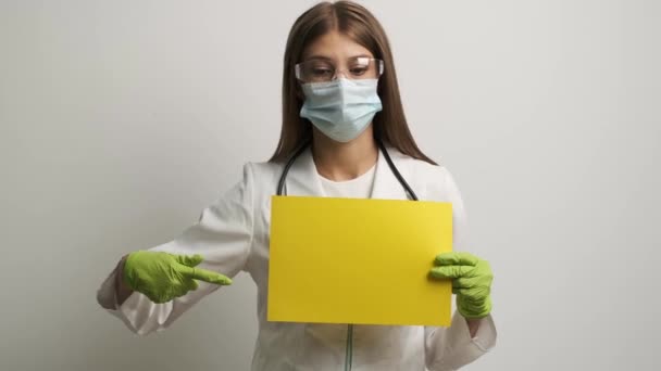 Femme médecin dans un masque d'apparence caucasienne tient dans ses mains une feuille A4 jaune — Video