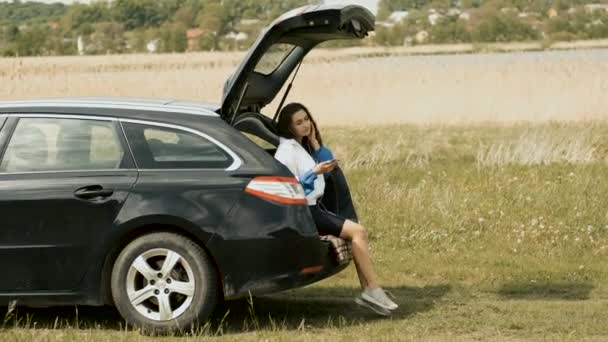 Happy girl sitting in the car in nature holding a phone in her hands and listening to music on headphones. Relax — Stock Video