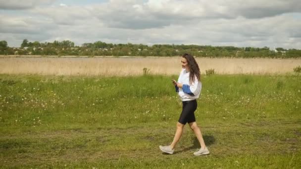 Mooi vrolijk meisje van Kaukasische verschijning wandelingen in het park kijkt naar de telefoon en luistert naar muziek op koptelefoon — Stockvideo