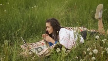 Neşeli, güzel Asyalı kız dizüstü bilgisayarla doğaya uzanıp ders çalışıyor.