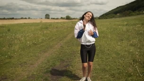 Beautiful happy girl in the park on the background of the lake listens to music on headphones and dances — Stock Video