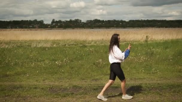 Mooi gelukkig lachend meisje wandelingen in het park op de achtergrond van het meer communiceert door middel van een video gesprek op een smartphone — Stockvideo