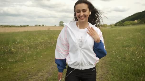 Mooi meisje van Kaukasische verschijning wandelingen in het park kijkt naar de telefoon en luistert naar muziek op koptelefoon — Stockvideo