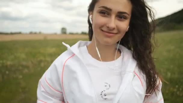 Belle fille d'apparence caucasienne marche dans le parc regarde le téléphone et écoute de la musique sur écouteurs — Video