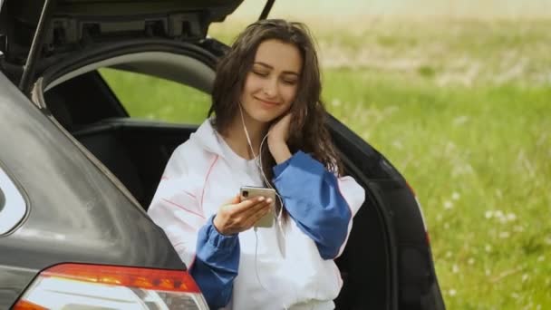 Doğada arabada oturan mutlu bir kız elinde telefonla kulaklıkla müzik dinliyor. Rahatla. — Stok video
