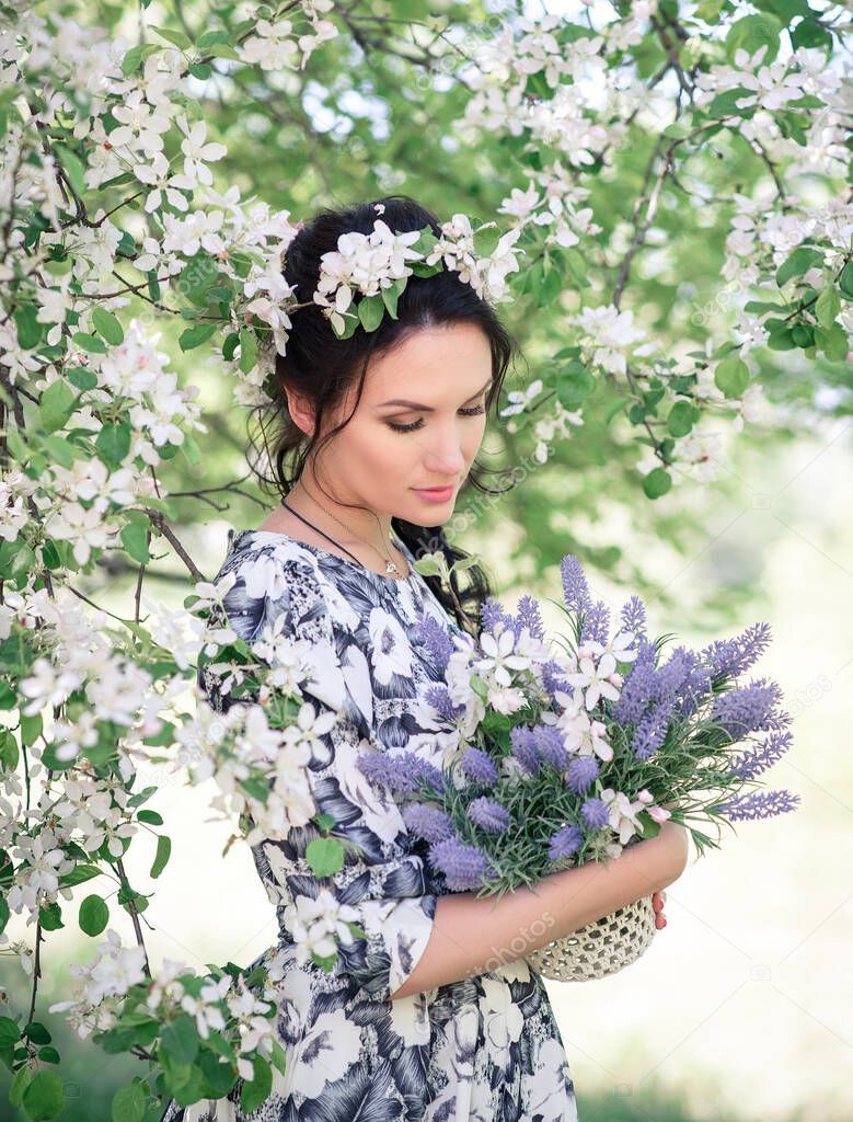 Girl with a bouquet of lilacs. A beautiful woman in a summer dress under a cherry blossom. Romantic lady with a wreath on her head in a blooming garden. Outdoor portrait