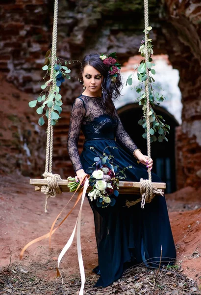 Mysterious woman in a black dress on a swing with flowers. Ruins of an old monastery. Black magic, Gothic beauty, mystical image. Halloween, Black widow