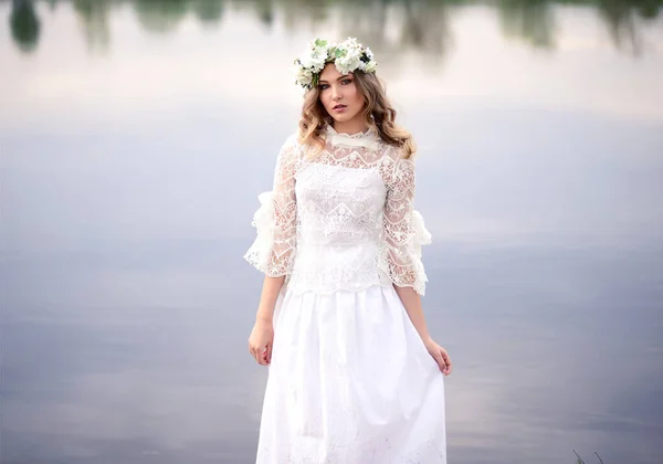 A beautiful girl in a white dress with a wreath of flowers stands near the river. Young girl on the background of a blue lake