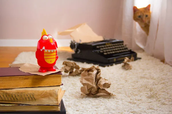 A red dragon toy stands on ancient books, and an out of focus old typewriter. White carpet, ginger cat and crumpled brown paper as background.