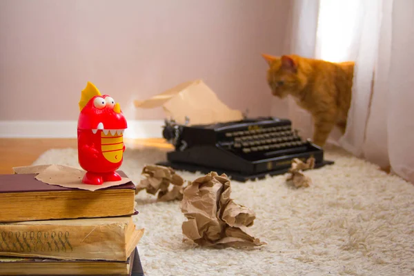 A red dragon toy stands on ancient books, and an out of focus old typewriter. White carpet, ginger cat became interested in the typewriter and crumpled brown paper as background.