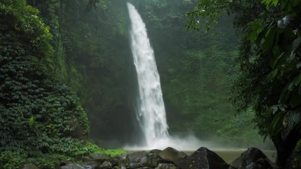 驚くべきヌンヌンの滝スローモーション落下水が水面を打つ緑豊かな葉のフレームの前に見えるいくつかの巨大な岩は風バリインドネシアから動いています — ストック動画