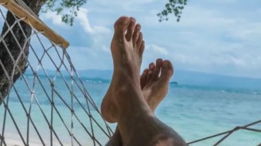 Sahilde çıplak ayakla sallanan beyaz kumlar, mavi okyanusun önünde tatilde bir hamakta sallanan.