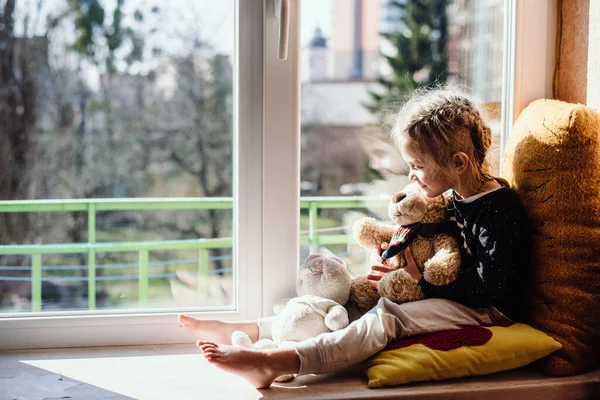 Cute little girl enjoying the sunshine while sitting at the window. The child looks out the window while sitting in the house. Vitamin D.  Film noise