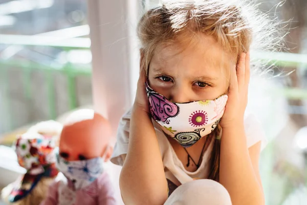 Koruyucu maske takan hasta bir çocuk pencerede oturur. Hasta enfeksiyonu önlemek için evde izole edilmiş. Coronavirus. Çocuğunuza covid-19, virüsler ve gribe karşı önleyici önlemler öğretmek.