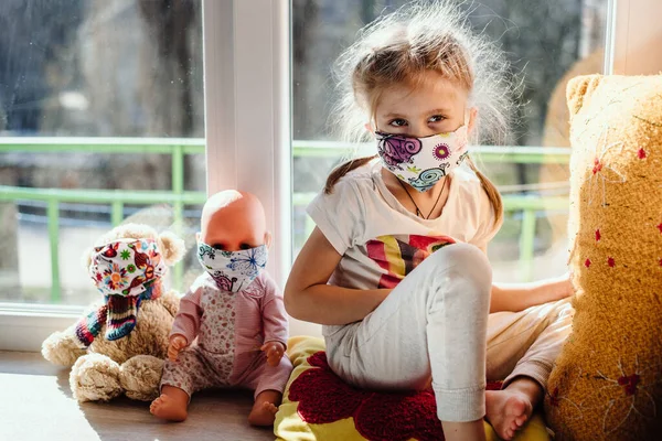 Teaching your child preventive measures against covid-19, viruses and flu. A baby girl in a protective mask puts a medical mask on her teddy bear toy. Care for loved ones. Basic hygiene rules. Film noise