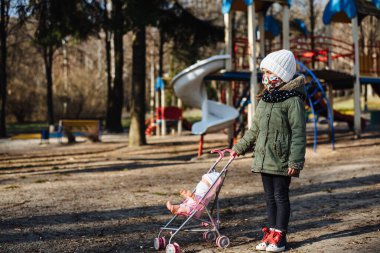 Kapalı oyun alanının arka planında koruyucu maskeli üzgün küçük kız. Tıbbi maskeli bir çocuk parkta yürüyor. COVID-19 'a karşı karantina önlemleri. Koronavirüs salgını.