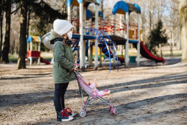 Kapalı oyun alanının arka planında koruyucu maskeli üzgün küçük kız. Tıbbi maskeli bir çocuk parkta yürüyor. COVID-19 'a karşı karantina önlemleri. Koronavirüs salgını.