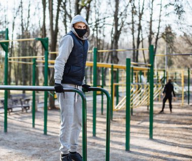 Çocuk parkında koruyucu maskeli ve eldivenli bir adam var. Gri kapüşonlu genç bir sporcu koronavirüs salgını sırasında parkta spor yapıyor. Ev aktivitelerinde kal. COVID-19.