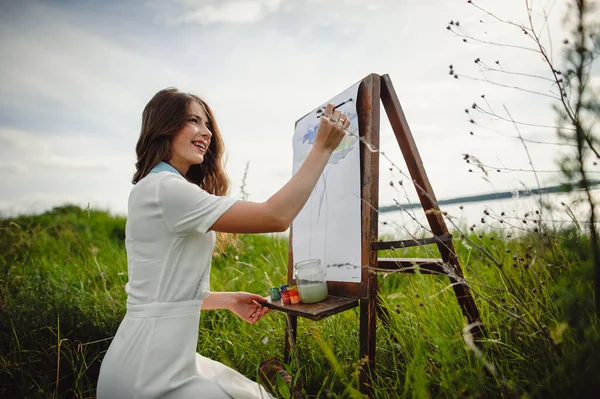 Easel with canvas, oil paint, palette and tools of the artist. Young artist paints a picture standing near an easel in nature. Girl painter artist is painting outdoors. Portrait of happy woman