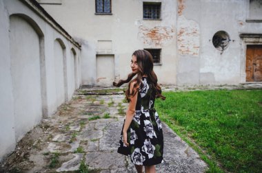 Elbiseli güzel esmer, eski kalenin avlusuna gider. Gri vintage binanın arka planında siyah elbiseli şık bir kız.