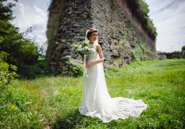 Beyaz elbiseli, güzel, şık bir gelin. Elinde düğün buketi taştan duvar arkasında poz veriyor. Gelin kalenin yakınında..
