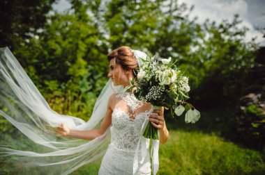 Gelinin ellerinde güzel bir buket beyaz çiçek ve yeşillikler. Tarz sahibi beyaz elbiseli bir gelin ve yeşil ağaçların arka planında bir peçe. Metin için yer.