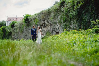 Beyaz elbiseli gelin ve smokinli damat eski kalenin taş duvarının yanında yeşil bir çayırla el ele tutuşuyorlar. Açık havada evlenen mutlu gelinler.