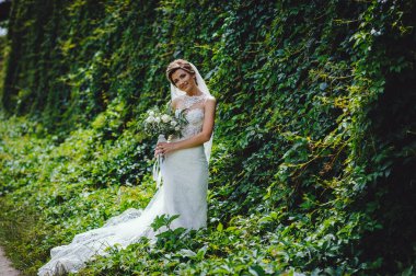 Lüks bir gelinlik içindeki güzel gelin yeşil doğal arka planda bir buket beyaz gül ve yeşillik tutuyor. Beyaz elbiseli mutlu gelinin portresi duvarın arkasında yeşilliklerle gülümsüyor..