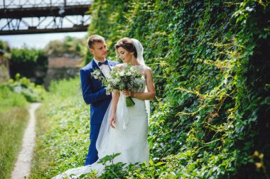Gelin ve damat yeşil doğanın arka planına karşı yüz yüze duruyorlar. Düğün gününde mutlu bir çift. Gelinin ellerinde muhteşem bir buket beyaz çiçek.