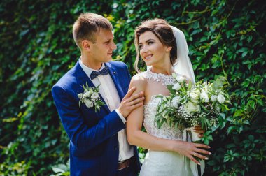 Birbirlerini seven güzel gelinlerin portresi. Gelin ve damat yeşil doğanın arka planına karşı yüz yüze duruyorlar. İnanılmaz gülümseyen bir düğün çifti. Güzel gelin ve şık damat.