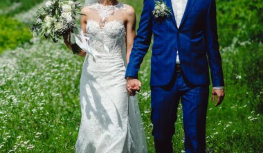 Smokinli, dantelli, damatlı beyaz elbiseli gelin yeşil çimen ve çiçeklerden oluşan bir arka planda el ele tutuşuyor. Düğün töreni açık havada..